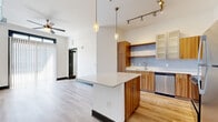 This single-floor unit showcases a thoughtfully designed open-concept layout with consistent light hardwood flooring connecting each area seamlessly. The kitchen serves as a focal point, featuring warm wood cabinetry paired with frosted glass-front uppers, a soft blue subway tile backsplash, and a full suite of stainless steel appliances including a range, microwave, dishwasher, and refrigerator. A central island with a white quartz-style countertop provides additional prep space and casual seating. Pendant lighting and a rail track system illuminate the kitchen, enhancing both function and style.

The living area benefits from high ceilings with exposed ductwork adding an industrial character, ceiling fans for comfort, and a large sliding glass door dressed in vertical blinds that floods the space with natural light and offers outdoor views. The bedroom areas feature clean white walls, dark baseboards, and ample outlet access. The bathroom is finished with large-format white floor tiles, a wood-toned vanity with an integrated sink, and a full-size tub and shower combo with classic white subway tile surround. The overall ambiance is clean, contemporary, and move-in ready.