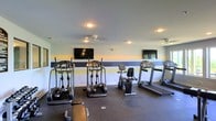 This single-floor fitness center offers a dedicated space for cardiovascular exercise and general wellness. The room is outfitted with a variety of cardio equipment, including elliptical trainers, treadmills, and a recumbent stationary bike, providing users with multiple options for their workout routines. Two wall-mounted flat-screen TVs are positioned throughout the room, allowing exercisers to stay entertained during their sessions. The flooring consists of durable rubber gym matting, which provides both safety and stability underfoot. Ceiling fans are installed overhead to promote airflow and comfort during workouts. The walls feature a bold horizontal striped accent in navy blue and golden yellow, adding a dynamic and motivating visual element to the space. Recessed lighting throughout the ceiling ensures the room is well-lit at all times. The overall layout is open and thoughtfully arranged, allowing for easy movement between equipment. The space maintains a clean, functional aesthetic that is well-suited for daily fitness use.