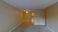 This single-floor space presents an open, functional layout with a clear visual connection between the main living area and the kitchen. The living area is finished with wall-to-wall carpet in a neutral tone, complemented by warm beige walls and white baseboards that run throughout, creating a clean and cohesive look. A ceiling fan with a built-in light fixture is centrally positioned, providing both air circulation and ambient lighting to the space.

The transition from the carpeted living area to the kitchen is marked by a shift to hard surface flooring, lending a practical distinction between the two zones. The kitchen, visible through the open layout, features white cabinetry and white appliances, including what appears to be a range and refrigerator. The cabinetry offers functional storage, and the overall kitchen design is straightforward and utilitarian.

The walls throughout maintain a consistent warm beige palette, contributing to a cohesive, understated ambiance. The space is unfurnished, allowing for a clear appreciation of the layout and the natural flow between areas. White trim and baseboards provide a crisp contrast to the walls, adding a degree of polish to the interior.