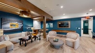 This thoughtfully designed single-floor communal space offers a versatile and inviting environment that blends lounge, dining, and recreation seamlessly. The interior is defined by striking deep teal accent walls that contrast beautifully against light, wide-plank wood-look flooring, creating a vibrant yet sophisticated atmosphere throughout.

The main living area features a comfortable seating arrangement with neutral-toned sofas and rounded armchairs centered around a sculptural coffee table. A slatted wood ceiling detail adds warmth and architectural interest overhead, while bold large-scale artwork adds personality to the walls. A wall-mounted flat-screen television anchors one side of the room, complemented by a full-size shuffleboard table for recreation.

A glass-paneled sliding partition elegantly separates the lounge from an adjoining dining and meeting area, allowing for visual connectivity while offering flexible privacy. The dining zone showcases a built-in banquette bench set against an artistic, textured feature wall, with pendant lighting casting a warm glow over teal velvet seating and communal tables.

Natural light filters in through large windows along the teal walls, enhancing the bright, open feel of the space. The overall ambiance is modern and social, balancing style with functionality to create a welcoming environment suited for both relaxation and gathering.
