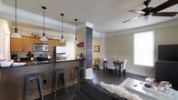 This two-floor space showcases a thoughtfully designed open-concept main level where the kitchen, dining, and living areas flow naturally into one another. The kitchen is well-appointed with stainless steel appliances, dark granite countertops, and warm wood cabinetry. A beadboard-paneled breakfast bar with industrial-style metal stools provides casual seating, while three pendant lights with dark fixtures hang overhead, adding both function and character. A distinctive structural column wrapped in chalkboard paint serves as a creative focal point, dividing the kitchen from the dining area while offering a customizable display surface. Crown molding runs throughout the main level, lending a polished, finished look to the space. Dark hardwood flooring anchors the entire open-concept area, providing a cohesive and grounded aesthetic. The dining area accommodates a full table with seating for multiple guests, while the adjoining living room features a comfortable sectional sofa. A window along the far wall allows natural light to brighten the space. The ceiling fan in the living area adds a practical element, contributing to overall comfort throughout the home.