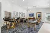 Located at 2235 Treehouse Ln in Corona, California, this single-floor community amenity space offers a well-appointed and thoughtfully designed gathering area. The layout flows seamlessly from a tiled entry foyer into a carpeted lounge and activity zone, creating distinct areas for socializing and recreation within a unified, open floor plan.

The lounge area is anchored by a neutral-toned sofa accented with blue throw pillows, a wall-mounted flat-screen TV, and a sleek mid-century modern media console. A full-length wood-framed mirror and a leather accent chair add warmth and character to the space. Large-scale abstract artwork adorns the soaring white walls, enhancing the sense of height and openness created by the elevated ceilings.

A foosball table and a round dining table with modern wire-frame chairs provide recreational and casual dining options. The entry corridor features large-format porcelain tile flooring, recessed lighting, and a pendant light fixture, transitioning smoothly into the carpeted main area with its multi-armed sputnik-style chandelier.

Natural light fills the space through multiple windows and glass doors, offering views of the outdoors. The overall design aesthetic is contemporary and inviting, combining clean white walls, layered area rugs, curated artwork, and modern lighting to create a vibrant yet comfortable community space.