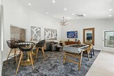 Located at 2235 Treehouse Ln in Corona, California, this single-floor community amenity space offers a well-appointed and thoughtfully designed gathering area. The layout flows seamlessly from a tiled entry foyer into a carpeted lounge and activity zone, creating distinct areas for socializing and recreation within a unified, open floor plan.

The lounge area is anchored by a neutral-toned sofa accented with blue throw pillows, a wall-mounted flat-screen TV, and a sleek mid-century modern media console. A full-length wood-framed mirror and a leather accent chair add warmth and character to the space. Large-scale abstract artwork adorns the soaring white walls, enhancing the sense of height and openness created by the elevated ceilings.

A foosball table and a round dining table with modern wire-frame chairs provide recreational and casual dining options. The entry corridor features large-format porcelain tile flooring, recessed lighting, and a pendant light fixture, transitioning smoothly into the carpeted main area with its multi-armed sputnik-style chandelier.

Natural light fills the space through multiple windows and glass doors, offering views of the outdoors. The overall design aesthetic is contemporary and inviting, combining clean white walls, layered area rugs, curated artwork, and modern lighting to create a vibrant yet comfortable community space.