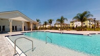 This community pool area in Port Orange, Florida, offers a well-appointed outdoor retreat designed for leisure and social enjoyment. The centerpiece is a generously sized swimming pool filled with clear, blue water, framed by elegant dark-tiled edging and accessible via stainless steel entry rails at multiple points. The pool's depth is suitable for recreational swimming, and posted pool rules ensure a safe environment for all users.

Surrounding the pool is a broad, neutral-toned paver deck that provides ample space for lounging and circulation. The pavers feature a refined stone-like finish that complements the community's upscale aesthetic. Tall, mature palm trees are strategically placed throughout the area, adding a tropical ambiance and natural shade.

Adjacent to the pool, a single-story clubhouse building with a classic coastal architectural style features a covered, columned pavilion equipped with a ceiling fan — offering a shaded outdoor gathering space that stays comfortable even on warm Florida days. Neatly maintained landscaping with flowering plants and mulched beds borders the structure.

The pool area is enclosed by a black wrought-iron fence, and views of the surrounding multi-story residential buildings are visible beyond the perimeter, reinforcing the sense of a vibrant, well-established community setting beneath sunny Florida skies.