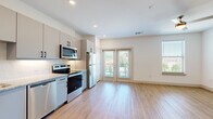 This thoughtfully designed single-floor unit offers a clean, modern aesthetic throughout, making excellent use of its compact footprint. Light wood-tone flooring flows consistently across all rooms, creating a cohesive and airy feel. The open-concept kitchen and living area serves as the heart of the space, where the kitchen is outfitted with sleek gray cabinetry featuring matte black hardware, granite-look countertops, a decorative tile backsplash, and a full suite of stainless steel appliances including a refrigerator, electric range, microwave, and dishwasher. The living area receives generous natural light through large windows and French doors that open to an outdoor balcony, enhancing the sense of openness. A ceiling fan adds both comfort and style to the living space. The hallway connects the main living areas to the bathroom and provides access to two closets, offering practical storage solutions. One closet houses a stacked in-unit washer and dryer, a highly convenient feature. The bathroom maintains the same modern design language, with gray vanity cabinetry, a granite-look countertop, matte black fixtures, and a wall-mounted storage cabinet. Recessed lighting and crisp white walls throughout contribute to the bright, contemporary ambiance of this well-appointed unit.
