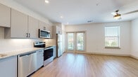 This thoughtfully designed single-floor unit offers a clean, modern aesthetic throughout, making excellent use of its compact footprint. Light wood-tone flooring flows consistently across all rooms, creating a cohesive and airy feel. The open-concept kitchen and living area serves as the heart of the space, where the kitchen is outfitted with sleek gray cabinetry featuring matte black hardware, granite-look countertops, a decorative tile backsplash, and a full suite of stainless steel appliances including a refrigerator, electric range, microwave, and dishwasher. The living area receives generous natural light through large windows and French doors that open to an outdoor balcony, enhancing the sense of openness. A ceiling fan adds both comfort and style to the living space. The hallway connects the main living areas to the bathroom and provides access to two closets, offering practical storage solutions. One closet houses a stacked in-unit washer and dryer, a highly convenient feature. The bathroom maintains the same modern design language, with gray vanity cabinetry, a granite-look countertop, matte black fixtures, and a wall-mounted storage cabinet. Recessed lighting and crisp white walls throughout contribute to the bright, contemporary ambiance of this well-appointed unit.
