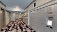 This mail room is a thoughtfully designed communal space built to handle the full range of mail and package needs for building residents. The room stretches into a corridor layout, with one wall lined by a large bank of secure parcel lockers finished in a clean, neutral tone — providing ample capacity for package deliveries of varying sizes. Along the opposite side, rows of individual resident mailboxes are neatly organized within decorative framed enclosures that feature a distinctive geometric cross-brace design, adding a subtle architectural character to an otherwise functional space. The flooring makes a strong visual statement with a bold, large-scale plaid or abstract patterned carpet in black, white, and brown tones, lending the space a modern and polished aesthetic. Recessed lighting in the ceiling provides consistent, even illumination throughout the corridor. A green-lit emergency exit sign is visible toward the far end of the room, and the neutral gray-toned walls keep the overall look clean and professional. The space is efficiently laid out to allow easy navigation and access to both mailboxes and parcel lockers, making it a practical and visually cohesive amenity within the building.