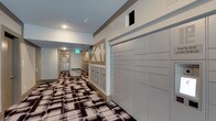 This mail room is a thoughtfully designed communal space built to handle the full range of mail and package needs for building residents. The room stretches into a corridor layout, with one wall lined by a large bank of secure parcel lockers finished in a clean, neutral tone — providing ample capacity for package deliveries of varying sizes. Along the opposite side, rows of individual resident mailboxes are neatly organized within decorative framed enclosures that feature a distinctive geometric cross-brace design, adding a subtle architectural character to an otherwise functional space. The flooring makes a strong visual statement with a bold, large-scale plaid or abstract patterned carpet in black, white, and brown tones, lending the space a modern and polished aesthetic. Recessed lighting in the ceiling provides consistent, even illumination throughout the corridor. A green-lit emergency exit sign is visible toward the far end of the room, and the neutral gray-toned walls keep the overall look clean and professional. The space is efficiently laid out to allow easy navigation and access to both mailboxes and parcel lockers, making it a practical and visually cohesive amenity within the building.
