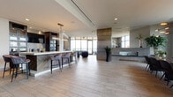 Located at 930 Speer Blvd in Denver, Colorado, this upscale amenity space delivers a seamless blend of contemporary design and elevated functionality. The open-plan layout is anchored by a well-appointed bar area featuring a striking marble-topped island with waterfall edges, dark cabinetry with brass hardware, integrated appliances, and a sculptural chandelier overhead. Light-toned tile flooring flows throughout, enhancing the bright, airy atmosphere.

A formal dining area accommodates large gatherings around an expansive long table, bathed in natural light from floor-to-ceiling windows with a modern multi-arm pendant fixture above. Adjacent lounge zones offer plush sectional seating, a built-in fireplace, and curated wall art, creating an inviting setting for relaxed conversation.

Floor-to-ceiling glass walls in the sitting area frame sweeping panoramic views of the Denver skyline and surrounding cityscape. A private outdoor terrace, accessed through sliding glass doors, extends the living space and provides an al fresco retreat with additional seating and open-air views. A herringbone wood accent wall adds warmth and texture throughout the interior. The overall ambiance is refined, modern, and thoughtfully designed for both social gatherings and intimate settings.