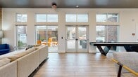 Located at 5025 Wembley Central Ln in Charlotte, NC, this single-floor community space offers a thoughtfully designed, open-concept layout that seamlessly integrates a kitchen, dining area, and living room. Soaring ceilings with a striking dark finish create a dramatic yet welcoming atmosphere throughout, while large floor-to-ceiling windows and sliding glass doors flood the interior with abundant natural light and provide direct views and access to an outdoor patio.

The kitchen is a standout feature, anchored by a substantial granite-topped island with geometric tile detailing and bar seating. Dark navy cabinetry, subway tile backsplash, and industrial-style pendant lighting give the kitchen a refined, contemporary character. Decorative signage referencing local Charlotte landmarks adds a distinctive neighborhood touch.

The adjacent dining area offers high-top tables and modern seating, ideal for casual gatherings. The living room is generously proportioned, highlighted by a textured brick accent wall mounting a large flat-screen television, warm wood-look tile flooring, and a comfortable sectional sofa arrangement. Glass doors connect directly to the outdoor patio, enhancing the sense of openness. A game table rounds out the lounge area, making this a versatile and inviting communal destination.