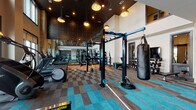 This contemporary fitness center is designed to support a wide range of workout styles within a single, thoughtfully laid-out space. The centerpiece of the facility is a soaring double-height ceiling that creates an open, energizing atmosphere, complemented by large floor-to-ceiling windows that flood the cardio area with abundant natural light and outdoor views. Stylish pendant lighting and a dark geometric accent ceiling add a modern, upscale character to the main training floor.

The main floor features a functional training zone anchored by a freestanding Octagon multi-rig, complete with TRX suspension straps, battle ropes, and a heavy punching bag for combat and functional fitness training. Adjacent to this is a dedicated free weights area with a full dumbbell rack, a flat bench, and a wall-mounted flat-screen television. A separate cardio zone houses treadmills, StairMasters, ellipticals, and a Concept2 RowERG rower, all positioned to take advantage of the natural light from the expansive windows.

A smaller, quieter alcove provides space for spin bikes and a rowing machine, alongside a water station and built-in shelving set against a textured tile feature wall. Throughout the facility, the flooring transitions between vibrant teal, warm orange, and neutral patterned carpet tiles, reinforcing the energetic yet polished design aesthetic. An upper-level mezzanine overlooks the main floor, adding to the grand sense of scale.