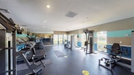 This single-floor fitness center offers a comprehensive workout environment designed to accommodate a variety of exercise routines. The space is defined by its open layout, dark rubber flooring, and a distinctive tri-color wall design featuring bands of tan, teal, and deep blue — giving the room a vibrant, energetic atmosphere.

Natural light floods the interior through large windows that line multiple walls, providing views of lush outdoor greenery and creating an inviting, airy feel. A full-length wall mirror expands the visual depth of the space and allows users to monitor their form during workouts.

The cardio section is well-stocked with multiple treadmills, elliptical trainers, and a stationary recumbent bike, all positioned to take advantage of the outdoor views. The strength training area includes a multi-function cable machine, a weight rack with medicine balls and dumbbells, and several resistance-based machines for targeted muscle training. Ceiling-mounted fans and a wall-mounted flat-screen television add comfort and entertainment to the workout experience.

Recessed lighting and HVAC vents in the textured ceiling ensure the space remains well-lit and climate-controlled throughout. Access to an adjacent outdoor area, likely a pool deck, is visible through glass doors, further enhancing the connectivity between the indoor fitness space and outdoor amenities.
