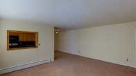 Located at 134 Innis Ave in Poughkeepsie, New York, this single-floor residence offers a straightforward and functional layout designed for comfortable everyday living. The main living area is generously sized, featuring wall-to-wall carpeting in a warm neutral tone that adds a soft, understated feel to the space. The walls are finished in a clean, light cream color, lending a bright and airy atmosphere throughout. A flush-mounted ceiling light fixture provides consistent overhead illumination, while baseboard heating units along the walls ensure efficient climate control during cooler months. A distinctive pass-through window connects the living area to the kitchen, featuring warm wood-toned cabinetry that adds a touch of natural character. This architectural detail promotes an open flow between the living and kitchen spaces while maintaining a degree of visual separation. Electrical outlets are conveniently positioned throughout the room, reflecting a practical and accessible design. The overall ambiance is clean, calm, and move-in ready, making it well-suited for those seeking a manageable, low-maintenance living space in the Poughkeepsie area.