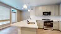 This well-designed single-floor residence offers a thoughtfully laid-out interior with a clean, contemporary aesthetic and warm, neutral tones throughout. Light wood-style flooring flows seamlessly across the entire space, creating a cohesive and inviting atmosphere.

The kitchen serves as the heart of the home, centered around a generous island topped with striking white marble-veined quartz countertops and an undermount stainless steel sink. Soft gray-toned shaker cabinets line the walls, providing ample storage, while a classic white subway tile backsplash adds a timeless touch. The kitchen is fully equipped with stainless steel appliances, including a gas range, over-the-range microwave, and a full-size refrigerator. A trio of modern geometric pendant lights hang above the island, adding both style and functional illumination. Natural light filters into the kitchen area through nearby windows, enhancing the open, airy feel.

The bathroom is clean and functional, featuring a floating white vanity with chrome hardware, a framed mirror, and a neutral stone-toned countertop. The space is finished with the same warm wood-style flooring found throughout the home.

A spacious walk-in closet offers multi-tiered shelving and hanging rods, providing generous storage and organizational flexibility. The floor plan, as seen in the dollhouse view, reflects a practical and well-proportioned layout designed for comfortable everyday living.