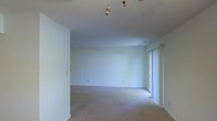 This single-floor residential space offers a clean, straightforward layout designed for comfortable everyday living. The main living area is finished with wall-to-wall carpeting in a neutral tone, providing a warm and quiet underfoot feel throughout. The walls are painted in a crisp, bright white, lending the space a fresh and open atmosphere that reflects light effectively and creates a sense of airiness.

Natural light enters the room through a full-height sliding glass door or large window panel positioned along one wall, allowing daylight to filter in and contributing to the overall brightness of the interior. A ceiling fan with a pull-chain mechanism is centrally mounted, offering both air circulation and a functional overhead fixture.

The room features standard electrical outlets distributed along the baseboards, ensuring practical usability throughout the space. Neutral-toned baseboard trim runs along the perimeter of the room, providing a clean transition between the walls and carpeted floor. The smooth, flat ceiling adds to the uncluttered aesthetic of the space.

Overall, this space presents a minimal, move-in-ready environment with a calm, neutral palette suited to a variety of furnishing styles. The open floor plan and natural light make it a versatile and welcoming living area.
