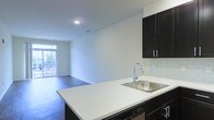 This single-floor space showcases an open-concept layout that seamlessly connects the kitchen and living area. The kitchen is outfitted with dark espresso cabinetry featuring sleek silver hardware, complemented by crisp white quartz-style countertops that extend into a peninsula with an undermount stainless steel sink and a contemporary pull-down faucet. A white subway tile backsplash adds a clean, classic detail to the kitchen walls, while integrated appliances, including a dishwasher, contribute to a streamlined aesthetic. The open layout allows for unobstructed sightlines into the living area, which features rich dark hardwood flooring that runs throughout the space. Neutral white walls keep the interior feeling bright and versatile. A window at the far end of the living area allows natural light to filter into the space, enhancing its airy atmosphere. Recessed lighting provides additional illumination throughout. The overall design reflects a modern, functional sensibility with a neutral palette well-suited to a variety of furnishing styles.