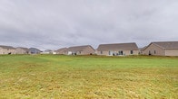 This single-story home presents a clean, understated exterior characterized by a neutral-toned stucco or siding finish beneath a gabled roofline. The structure is set well back from the front of the property, allowing for a broad, open lawn that stretches expansively in front of and around the home. The generous green yard provides ample outdoor space, offering a sense of openness and room to move. At the rear of the home, a patio area is visible, suggesting a designated outdoor living or entertaining space that extends the usability of the property beyond its interior. Multiple windows punctuate the facade, allowing for natural light to enter the home and providing views out to the surrounding landscape. The property sits within a residential neighborhood, with neighboring homes of similar architectural style visible nearby. The overall setting conveys a quiet, suburban character with plenty of open sky and green space. The single-floor layout offers convenient, accessible living with all areas on one level, making navigation throughout the home straightforward and practical. The combination of a well-maintained yard, patio space, and unobstructed surroundings gives this property a comfortable, functional, and approachable character suited for everyday living.