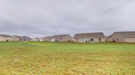 This single-story home presents a clean, understated exterior characterized by a neutral-toned stucco or siding finish beneath a gabled roofline. The structure is set well back from the front of the property, allowing for a broad, open lawn that stretches expansively in front of and around the home. The generous green yard provides ample outdoor space, offering a sense of openness and room to move. At the rear of the home, a patio area is visible, suggesting a designated outdoor living or entertaining space that extends the usability of the property beyond its interior. Multiple windows punctuate the facade, allowing for natural light to enter the home and providing views out to the surrounding landscape. The property sits within a residential neighborhood, with neighboring homes of similar architectural style visible nearby. The overall setting conveys a quiet, suburban character with plenty of open sky and green space. The single-floor layout offers convenient, accessible living with all areas on one level, making navigation throughout the home straightforward and practical. The combination of a well-maintained yard, patio space, and unobstructed surroundings gives this property a comfortable, functional, and approachable character suited for everyday living.