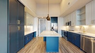 This single-floor space showcases a thoughtfully designed kitchen as its centerpiece, combining bold navy blue lower cabinetry with soft light grey upper cabinets, all accented by brushed gold hardware. A generously sized central island features a polished quartz countertop with subtle veining, complemented by a trio of antique brass pendant lights suspended overhead. The kitchen is well-equipped with stainless steel appliances, including a French door refrigerator, range with microwave, and dishwasher. A herringbone-patterned tile backsplash adds texture and visual interest, while under-cabinet lighting illuminates the workspace below. Wood-look flooring runs throughout, adding warmth and continuity to the space. Natural light filters in through tall windows and a glass-paneled exterior door, enhancing the open, airy atmosphere. The bathroom continues the refined aesthetic with grey tile walls and flooring, a marble-topped vanity with white cabinetry and brass fixtures, a tub-shower combination with iridescent subway tile surround, and elegant wall sconce lighting. The overall design balances contemporary style with classic detailing.