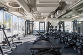 This fitness center offers a well-appointed, elongated layout designed to accommodate a wide variety of workout routines. The space is defined by its floor-to-ceiling glass curtain walls along one side, flooding the interior with abundant natural light and providing sweeping views of the surrounding urban skyline. The open, industrial-style ceiling features exposed ductwork, white-painted mechanical systems, and distinctive circular pendant light fixtures that contribute to the facility's contemporary aesthetic.

The flooring throughout consists of durable, large-format rubber tiles in grey tones, providing both safety and functionality. Cardio equipment is prominently featured, with a full row of modern treadmills and elliptical trainers positioned along the window-facing side to take advantage of the views. Strength and resistance machines by HOIST and other brands are arranged along the interior wall, offering a comprehensive range of targeted muscle training options. Additional equipment, including rowing machines and stationary bikes, is distributed throughout the space.

The interior walls are finished in a neutral grey and white palette, complemented by accent elements such as concrete columns and built-in cabinetry. Several private rooms with glass-paneled doors are accessible from the main floor, and a water station is conveniently located within the facility. Overall, the space maintains a clean, professional atmosphere suited for serious fitness use.