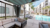 Located at 200 W Pomona Ave in Monrovia, California, this outdoor cabana space offers a well-appointed poolside retreat within a modern residential community. The single-level space is defined by a pergola-style retractable canopy with rich teal fabric panels that provide generous shade while maintaining an open, airy atmosphere. Draping privacy curtains frame the perimeter, allowing the space to be enclosed or opened to the surrounding environment as desired.

The seating area is furnished with wicker sectional sofas accented by striped and solid throw pillows, creating a comfortable and stylish lounge environment. A standout architectural feature is the built-in glass-front fireplace embedded in the adjacent wall, filled with glass beads that add a contemporary decorative element and provide warmth during cooler evenings.

From the cabana, views extend directly to the sparkling pool, lined with a row of chaise lounges ideal for sunbathing. Palm trees and lush plantings add a resort-like ambiance to the surroundings. The broader community backdrop features a modern multi-story building with balconies and clean white facades. The overall atmosphere is relaxed and upscale, making this space well-suited for social gatherings or quiet outdoor relaxation in the Southern California climate.