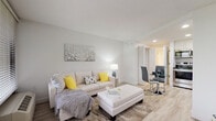 This single-floor unit offers a clean, contemporary design throughout. The open-concept living and dining area is anchored by light wood-look flooring, neutral white walls, and a textured ceiling. A large sliding glass door and wide window frame views of lush greenery, filling the space with natural light. The living area comfortably accommodates a sectional sofa centered around an upholstered ottoman, while a glass-top dining table and modern chairs are positioned nearby, delineated by louvered partition doors that add a subtle architectural detail.

The kitchen flows directly from the dining area and features crisp white cabinetry extending to the ceiling, white quartz-style countertops, and stainless steel appliances including an electric range and over-the-range microwave. The layout is efficient and well-organized, maximizing storage throughout.

The bathroom continues the bright, modern aesthetic with large-format marble-look porcelain floor tiles, white shaker-style cabinetry with brushed nickel hardware, and a generous vanity with ample counter space and drawer storage. Recessed lighting enhances the clean, polished finish. Overall, the unit presents a cohesive, well-appointed design with a bright and welcoming ambiance.