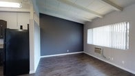 This single-floor space showcases a thoughtfully designed interior that blends modern aesthetics with functional details. The room features striking exposed white-painted wooden beams across the ceiling, adding architectural character and a sense of open volume overhead. Underfoot, dark hardwood-style flooring runs throughout, providing a warm yet contemporary foundation that complements the overall design palette. A bold charcoal accent wall serves as a dramatic focal point, offering a strong visual contrast against the lighter tones of the surrounding walls, trim, and ceiling. Natural light enters through a window fitted with vertical blinds, brightening the space while allowing for privacy and light control. A wall-mounted air conditioning unit ensures year-round comfort. The clean white baseboards and trim provide crisp definition throughout the room, tying together the neutral and dark tones cohesively. The overall ambiance is modern and understated, with an emphasis on clean lines and a balanced contrast between light and dark elements, making it a versatile space suitable for a variety of uses.