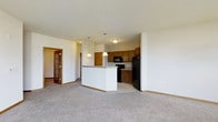 This single-floor unit is designed with an open-concept layout that seamlessly connects the living area and kitchen, creating a cohesive and functional space. The living area features neutral-toned carpet that adds warmth and comfort underfoot, while the smooth white walls and light-colored ceilings contribute to a bright, airy ambiance throughout.

The kitchen is positioned at the heart of the unit and is outfitted with warm medium-toned wood cabinetry that provides ample storage. A central peninsula serves as a natural dividing point between the kitchen and living space, topped with a stone-style surface and illuminated by two pendant lights that hang from the ceiling, adding both style and practical task lighting. Black appliances, including a refrigerator, range, and over-the-range microwave, complement the cabinetry for a cohesive look. The kitchen floor transitions to a hard surface, distinguishing it clearly from the carpeted living area.

A doorway visible on the left side of the space, framed by a distinctive wood-trimmed door with decorative glass paneling, leads to an additional room, adding to the unit's overall functionality and flow.