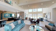 This single-floor clubhouse presents an expansive, open-concept layout designed for comfort and community gathering. The space is anchored by dramatically high ceilings that amplify the sense of openness, complemented by an abundance of natural light streaming through large floor-to-ceiling windows along the back wall, which frame views of the surrounding outdoor landscape.

The interior is finished with light hardwood-style flooring and neutral wall tones, creating a clean, contemporary backdrop. A statement chandelier with a modern globe design serves as a focal point overhead, while additional pendant lighting illuminates a bar or counter area along the left side of the space—ideal for social gatherings or events.

The lounge area is thoughtfully arranged with multiple seating zones, including a vibrant teal tufted sofa, dark upholstered armchairs, and glass-top accent tables, blending bold color accents with sophisticated neutrality. The open layout transitions seamlessly from the lounge area to the bar and additional seating near the windows, fostering a welcoming, social atmosphere throughout.