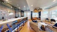 This thoughtfully designed communal lounge and co-working space occupies a single floor, offering a well-appointed environment that seamlessly blends industrial character with contemporary style. The open-concept layout is anchored by a long, marble-topped bar counter lined with modern wood-and-blue-metal barstools, set against a striking exposed brick wall framed by dark steel-framed glass partitions — a nod to industrial design sensibilities.

The lounge area is warm and inviting, centered around a rich cognac leather sofa, upholstered accent chairs, and a patterned area rug. A chess set rests on a low wooden coffee table, adding a social, recreational touch. Overhead, oversized woven pendant lights cast a warm glow throughout the space, complementing the recessed lighting embedded in the two-tone ceiling — a combination of dark charcoal and crisp white.

Light oak hardwood flooring flows throughout, tying together the various functional zones. Multiple windows along the perimeter flood the space with natural light and offer views of the surrounding neighborhood. Built-in navy blue bookshelves frame the windows, adding depth and a curated aesthetic.

A compact kitchenette features sleek gray cabinetry, open shelving, a coffee station, and a mini refrigerator — ideal for everyday convenience. A wall-mounted flat-screen TV and dedicated desk area round out the space, making it equally suited for relaxation and productivity.