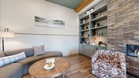 This single-floor combined living and dining space in Richmond, TX, is thoughtfully designed to maximize both comfort and functionality. The living area features warm hardwood-style flooring throughout, complemented by elevated ceilings with exposed wood beam accents and a teal-painted ceiling detail that adds a distinctive architectural touch. A floor-to-ceiling brick fireplace serves as a striking focal point, flanked by a custom built-in bookcase finished in a muted sage-gray tone, complete with integrated lower cabinetry and display shelving. Seating includes a curved upholstered sofa and a floral accent chair, centered around a round reclaimed wood coffee table. Elegant French doors with geometric patterned glass panels provide entry to the space, connecting it to adjacent areas while allowing visual openness. A large window with a roller shade allows natural light to filter in and offers a view of an outdoor seating area. The dining area occupies a dedicated corner, accommodating a round dark-topped table surrounded by cane-back chairs beneath framed artwork. A consistent wainscoting-style stripe runs along the walls, unifying the overall aesthetic and creating a polished, cohesive ambiance throughout.