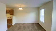 This single-floor space presents a straightforward and functional layout centered around an open living and dining area. The room is finished with wall-to-wall carpet in a neutral beige tone, contributing to a quiet and comfortable atmosphere underfoot. Walls are painted in a clean off-white, creating a bright and open feel throughout. A pendant light fixture hangs from the ceiling, providing focused overhead illumination to the dining area and serving as a subtle design accent within the otherwise understated space. Natural light enters through a window fitted with horizontal blinds on the right-hand wall, offering ventilation and an exterior view. An HVAC vent is visible in the ceiling, indicating a centralized climate control system. Adjacent to the living and dining area, a partial view of the kitchen is visible, revealing light wood cabinetry and a countertop pass-through or bar-height ledge that connects the two spaces. The textured ceiling adds a modest architectural detail consistent with residential construction. Overall, the space offers a clean, move-in-ready environment with a neutral palette well-suited to a variety of interior styles and furnishing choices.