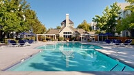 Located in South Jordan, Utah, this outdoor amenity space centers around a generously sized swimming pool with crystal-clear blue water, designed to accommodate active recreation and relaxed leisure alike. The pool features multiple entry points, including steps and handrails, ensuring easy access throughout. Surrounding the pool, a broad concrete sun deck provides ample space for lounging, furnished with rows of navy blue chaise lounge chairs paired with coordinating scalloped umbrellas for shaded comfort. A modern pergola structure with slatted wood screening offers an additional shaded retreat and casual gathering area along the deck's perimeter. Mature deciduous trees are thoughtfully integrated throughout the space, providing natural shade and a sense of greenery that softens the hardscape. Colorful seasonal plantings and well-maintained shrubs add vibrancy along the walkways and borders. A charming clubhouse building anchors one end of the pool area, featuring a distinctive peaked roofline, a chimney, and large windows that open toward the deck, suggesting interior amenity spaces beyond. The overall ambiance is resort-like, combining a classic architectural aesthetic with practical outdoor amenities, all set beneath the open skies of Utah's sunny climate.