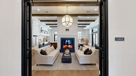 Located at 601 Gilcrest Farm Rd in Wake Forest, NC, this thoughtfully designed single-floor space spans an impressive footprint and delivers a refined blend of modern farmhouse and contemporary aesthetics. Wide-plank hardwood flooring flows throughout, unifying the open layout.

The living room is a showpiece, anchored by a sleek fireplace with a navy blue surround and flanked by dual sofas beneath exposed dark wood ceiling beams and a geometric pendant light. Black-framed windows flood the space with natural light and offer views of the outdoor pool area.

The dining room commands attention with a dramatic dark ceiling, a cascading crystal chandelier, and a long communal table bordered by leather and woven chairs. Deep navy cabinetry lines one wall, while floor-to-ceiling black-framed windows invite abundant daylight.

A cozy lounge nook features sage green millwork with upholstered booth seating — ideal for intimate gatherings. The hallway entry is punctuated by striking steel-and-glass French doors with a transom window above.

The spacious primary bedroom offers a serene retreat with tall windows, flowing drapes, and rich gray walls. A standout feature is the dedicated golf simulator room, equipped with a full-swing screen, turf mat, and integrated technology — a rare and impressive amenity.