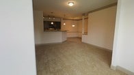 This single-floor residential space showcases an open-concept design that seamlessly connects the living area and kitchen. The living area is finished with neutral-toned wall-to-wall carpeting that lends warmth and comfort underfoot, while the open layout creates a spacious, airy feel throughout.

The kitchen is positioned at the rear of the main living space and features dark cabinetry that provides a rich contrast against the light-toned walls. A raised breakfast bar serves as both a functional workspace and a natural divider between the kitchen and living area. Stainless steel and black appliances, including a built-in microwave, add a modern touch to the culinary space.

Pendant lighting is a standout design element, with multiple hanging fixtures illuminating both the kitchen and an adjacent dining nook area. A decorative column accent adds an architectural detail that contributes character to the open floor plan. The walls are finished in a soft, neutral palette, and a subtle chair rail detail runs along the perimeter, adding a classic touch to the overall aesthetic. Flush-mount ceiling lighting in the main living space ensures the area is well-lit throughout. The overall ambiance is clean, neutral, and move-in ready.