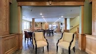 This well-appointed single-floor space offers an open, flowing layout designed for comfort and versatility. The interior is anchored by rich wood architectural details, including decorative columns with carved capitals and warm wood trim that frame the ceiling and doorways, lending a classic, refined character throughout.

The stone tile flooring runs continuously across the expansive open floor plan, unifying the multiple functional areas. Recessed lighting is abundant across the drop ceiling, ensuring the space is well-lit and inviting at all times of day. Natural light also filters in through windows along the perimeter, further brightening the warm, golden-toned walls.

The layout accommodates several distinct seating arrangements. Near the entrance, a pair of elegant cream upholstered armchairs with dark wood frames creates a welcoming reception area. Beyond, a bar-height counter with dark stools suggests a casual dining or social hub. Further into the space, additional dining and lounge areas are visible, separated by the decorative columns that define zones without closing off the open feel.

A staircase is visible toward the rear, and what appears to be a kitchen or service area is nestled in the background. The overall ambiance is warm, polished, and functional — well-suited as a community lounge, clubhouse, or upscale gathering venue.