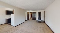 This single-floor residence offers a practical and functional layout designed to maximize living space. The open-concept living room is generously sized, featuring neutral wall-to-wall carpeting, a textured ceiling, and an abundance of natural light provided by large vertical-blind-covered sliding glass doors that lead to an outdoor balcony. An additional window further brightens the space, creating an airy atmosphere throughout.

The kitchen is positioned adjacent to the living area and features a classic galley-style configuration with dark espresso-toned cabinetry lining both walls, offering ample upper and lower storage. White appliances, including a full-size refrigerator and dishwasher, complement the light countertops, while dark hardwood-style flooring provides a clean contrast to the cabinetry.

The bedroom is carpeted and well-proportioned, equipped with a ceiling fan and light fixture. Multiple closet doors provide practical storage options. The bathroom includes a full tub and shower combination with neutral tile surround, a toilet, and dark-toned flooring. Throughout the unit, dark wood-toned doors and trim provide a consistent, cohesive design element against the neutral white walls.