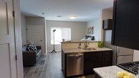 This single-floor space showcases an open-concept layout that seamlessly connects the kitchen, living, and sleeping areas into one cohesive environment. The kitchen serves as a central feature, equipped with granite countertops, dark espresso cabinetry, a stainless steel undermount sink, and a stainless steel dishwasher. The kitchen island provides additional prep and storage space, while the layout allows for easy interaction between the cooking and living zones.

The flooring throughout is a warm-toned hardwood-style material that lends consistency and a modern aesthetic across all areas. Neutral gray walls complement the overall palette, creating a clean and contemporary feel. A window fitted with horizontal blinds allows natural light to filter into the space, brightening the central area shared by the living and sleeping zones.

The living area accommodates a sofa and area rug, offering a defined relaxation zone, while the sleeping area is visible in the background, benefiting from the same open, airy atmosphere. A white paneled door provides access to an additional room or storage space. The overall design prioritizes functionality and an uncluttered, modern aesthetic throughout.