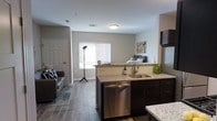 This single-floor space showcases an open-concept layout that seamlessly connects the kitchen, living, and sleeping areas into one cohesive environment. The kitchen serves as a central feature, equipped with granite countertops, dark espresso cabinetry, a stainless steel undermount sink, and a stainless steel dishwasher. The kitchen island provides additional prep and storage space, while the layout allows for easy interaction between the cooking and living zones.

The flooring throughout is a warm-toned hardwood-style material that lends consistency and a modern aesthetic across all areas. Neutral gray walls complement the overall palette, creating a clean and contemporary feel. A window fitted with horizontal blinds allows natural light to filter into the space, brightening the central area shared by the living and sleeping zones.

The living area accommodates a sofa and area rug, offering a defined relaxation zone, while the sleeping area is visible in the background, benefiting from the same open, airy atmosphere. A white paneled door provides access to an additional room or storage space. The overall design prioritizes functionality and an uncluttered, modern aesthetic throughout.