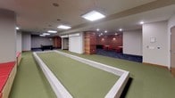 This community room is a generously sized, single-floor recreational space designed to accommodate a wide variety of social and leisure activities. The room is divided into two distinct zones distinguished by their flooring: a green turf area and a dark blue carpeted section. The turf zone houses a multi-hole indoor putting green with contoured surfaces, a full-length bocce ball court framed by low concrete borders, and built-in bench seating with red upholstered cushions and natural wood bases. A foosball table and wall-mounted flat-screen television add to the entertainment options in this area. A large interior window overlooks what appears to be a fitness room, adding visual connectivity between spaces. The carpeted zone transitions into a more lounge-like setting, featuring a pool table, round tables with red chairs for casual gathering, and a bar area accented with warm wood paneling, pendant lighting, and bar stool seating. A roll-up service window provides access to an adjacent storage or service area. Throughout the room, flush and suspended panel lighting provides consistent, even illumination. The neutral gray walls and warm wood trim details create a clean, welcoming ambiance suited for community gatherings and recreation.