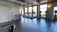 This contemporary fitness center is designed to provide a motivating and functional workout environment. The space is anchored by a row of treadmills and elliptical machines positioned along expansive floor-to-ceiling windows, offering open views of the outdoors and an abundance of natural light that creates an energizing atmosphere throughout the day. The flooring consists of sleek, dark hardwood-style planks that lend the space a polished, modern aesthetic while remaining durable underfoot. A dedicated free weights area lines one wall, featuring a well-organized rack of color-coded kettlebells in a range of sizes, along with additional functional training accessories. Full-length mirrors along the adjacent wall enhance the sense of space and allow for proper form monitoring during workouts. The ceiling is finished with a structured coffer design accented by recessed lighting, complemented by industrial-style ceiling fans that ensure proper air circulation. The overall design balances clean, neutral tones — including gray walls and dark flooring — with bright, open sightlines, resulting in a professional and inviting space suitable for a wide range of fitness activities.
