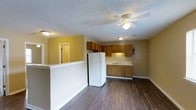 Located at 3250 Boardwalk Dr in Greenville, NC, this single-floor unit offers a practical and comfortable living layout. The open-concept design connects the main living area to the kitchen, creating a spacious and functional flow throughout the space. The kitchen is equipped with warm oak cabinetry, a white refrigerator, and a double-basin sink set beneath upper and lower cabinets that provide ample storage. Light-colored countertops complement the natural wood tones of the cabinetry. The living area features rich, dark hardwood-style flooring that runs throughout, adding warmth and visual continuity to the space. Walls are painted in a warm yellow tone, contributing to a bright and inviting ambiance. A ceiling fan with a built-in light fixture is centrally positioned, ensuring good air circulation and illumination. The textured popcorn ceiling and clean white trim provide a classic finish. A half-wall partition subtly defines the transition between the living and kitchen areas. A doorway visible from the main space suggests additional rooms beyond, offering further privacy and utility. Overall, this unit provides a well-organized, cozy living environment well-suited to everyday comfort.