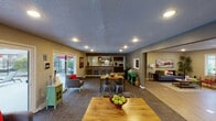 This single-floor community clubhouse offers a thoughtfully designed layout that blends functionality with a warm, inviting ambiance. The entrance greets visitors with a bright lounge area featuring hardwood-style flooring, a bold red door with frosted glass panels, a plush blue sofa, and curated wall art that sets a lively, contemporary tone.

Beyond the entry, the space opens into a spacious multi-purpose gathering area with textured ceilings and recessed lighting throughout. This central zone is furnished with multiple dining tables and metal industrial-style chairs, making it ideal for social gatherings and community events. Large sliding glass doors and windows along the perimeter flood the space with natural light and provide views of an outdoor patio and pool area.

A full kitchen sits at the rear of the main gathering space, outfitted with dark wood cabinetry, stainless steel appliances including a refrigerator and range, and pendant lighting overhead. A wraparound bar counter with rustic wooden stools offers additional casual seating.

Adjacent areas include a dedicated office or administrative space with dark wood desks, and a secondary seating and activity area. A windowed fitness center is visible from one of the interior rooms, equipped with cardio machines and free weights. The cohesive color palette of gray, tan, and accents of red and green ties the entire space together seamlessly.