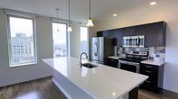 This contemporary kitchen is defined by clean lines and a striking contrast between its crisp white surfaces and bold dark cabinetry. The centerpiece is a generous waterfall-edge island finished in bright white, equipped with an undermount sink and a sleek arc faucet, offering ample prep and gathering space. Pendant lighting hangs above the island in a trio of varying heights, providing both task lighting and a stylish design accent.

The kitchen is outfitted with a full suite of stainless steel appliances, including a French door refrigerator, a built-in microwave, and an electric range, all set against dark flat-panel upper and lower cabinetry that extends to the ceiling for maximum storage. A subtle tile backsplash adds texture behind the cooking area.

Two floor-to-ceiling windows flood the space with natural light and frame expansive city views, creating an open, airy atmosphere. Dark hardwood-style flooring grounds the space and complements the cabinetry, lending a cohesive, modern aesthetic throughout. The overall ambiance is polished, functional, and well-suited for both everyday living and entertaining.
