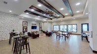 Located at 231 Noblewood Dr in San Antonio, TX, this single-story community clubhouse presents a thoughtfully designed gathering space that blends rustic charm with modern comfort. The open-plan interior is anchored by soaring ceilings adorned with bold, dark-stained exposed wood beams and industrial-style windmill ceiling fans, creating a striking architectural focal point overhead. Underfoot, a herringbone-patterned hardwood-style floor flows throughout, lending warmth and visual continuity across the entire space.

The lounge area features rich leather armchairs and sofas arranged around a round live-edge coffee table, set atop a vibrant patterned area rug. A shiplap accent wall serves as a media backdrop, flanked by framed botanical butterfly artwork, while a stacked stone column adds a natural, textural contrast. The dining area offers multiple wood tables and chairs bathed in natural light streaming through large, dark-framed windows that overlook an outdoor courtyard and patio.

The fully equipped kitchen includes dark wood cabinetry, white quartz countertops, a hexagonal tile backsplash, a stainless steel double wall oven, and a French door refrigerator, making it well-suited for community events and gatherings. The overall ambiance is warm, inviting, and community-focused.