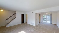 This two-story home offers three bedrooms and one and a half bathrooms across a well-proportioned floor plan. The main level features an open living area finished with light-toned carpet and bright white walls, creating a clean, neutral backdrop throughout. A staircase along the side wall provides access to the upper floor, where the bedrooms are likely located. On the main level, a half bathroom is accessible via a dark-wood door off the living area, adding everyday convenience. The layout opens toward the rear of the home, where a sliding glass door or window brings in natural light and provides a view of and access to an outdoor area, enhancing the sense of openness. A closet with louvered doors is visible near the transition to the rear portion of the home, offering practical storage. The kitchen is positioned at the back, with a refrigerator visible and natural light filtering in from outside. HVAC vents are present both on the walls and floors, ensuring climate control throughout. The overall design is straightforward and functional, with a neutral palette that provides a versatile foundation for personalization.