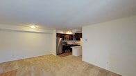 This single-floor unit features an open-concept layout that seamlessly connects the living area to the kitchen. The spacious living area is finished with neutral-toned wall-to-wall carpeting, creating a warm and comfortable atmosphere. Crisp white walls and a flush ceiling light fixture contribute to a clean, bright interior.

The kitchen is well-appointed with rich, dark wood cabinetry that provides ample storage. Stainless steel appliances — including a full-size refrigerator, electric range, and built-in microwave — offer a modern, functional aesthetic. Track lighting illuminates the workspace, while a peninsula counter with a breakfast bar serves as both a practical prep area and a natural dividing point between the kitchen and living space. The kitchen floor transitions to a hard surface, distinguishing it from the carpeted living area.

A wall-mounted thermostat is visible near the kitchen, indicating individual climate control. The open layout and neutral color palette create a versatile environment that adapts easily to a variety of furnishing styles, making this a practical and well-designed single-level living space.