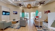This single-story clubhouse embraces a vibrant coastal aesthetic throughout its well-appointed interior. The main lounge area immediately captures attention with its soaring coffered ceilings adorned with white-painted beams, ceiling fans, and a distinctive woven-sphere chandelier. Full-size surfboards and paddleboards mounted as wall art reinforce the beach-inspired theme, while light wood-look flooring, ocean-toned area rugs, and neutral upholstered seating create a welcoming atmosphere. A wall-mounted television and a built-in fireplace anchor one side of the lounge, and French doors along the far wall provide direct views and access to an outdoor pool area, flooding the space with natural light.

Adjacent to the main lounge, a dedicated seating area features a reception desk, additional comfortable furnishings, and refined coastal accents. A bright dining nook with soft blue walls, a coffered ceiling, and large windows offers seating at dark wood tables paired with cream upholstered chairs.

The kitchenette presents a light-filled, functional layout with crisp white cabinetry, a mosaic tile backsplash, a quartz countertop island, a beverage refrigerator, and a coffee station, all accented by bold gold pendant lighting and soft aqua walls—completing the space's cohesive, resort-inspired character.