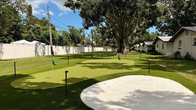Encantadora vista al aire libre con un putting green bien cuidado rodeado de exuberante vegetación y pintorescas cabañas bajo un cielo azul claro. - Waterside Village