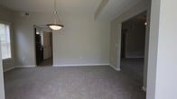 This two-floor space offers a straightforward and functional layout designed with clean lines and a neutral aesthetic throughout. The main living and dining area is an open, combined space finished with plush wall-to-wall carpeting in a warm gray tone, creating a comfortable underfoot feel. Neutral gray walls provide a versatile backdrop that adapts well to a variety of furnishing styles and décor choices.

A standout feature of this area is the classic bowl-style pendant light suspended from the ceiling by a chain, serving as a central focal point while providing ambient overhead lighting. The textured ceiling adds subtle visual interest to the space. Structural beam detailing is visible overhead, contributing to the architectural character of the room.

The open layout connects naturally to adjacent spaces, with doorways visible leading to additional rooms including what appears to be a kitchen area with dark appliances to one side and a hallway corridor leading further into the unit. White panel doors and crisp white baseboards offer clean contrast against the softer tones of the walls and flooring. Overall, the space presents a clean, move-in ready environment with a calm, neutral ambiance throughout.